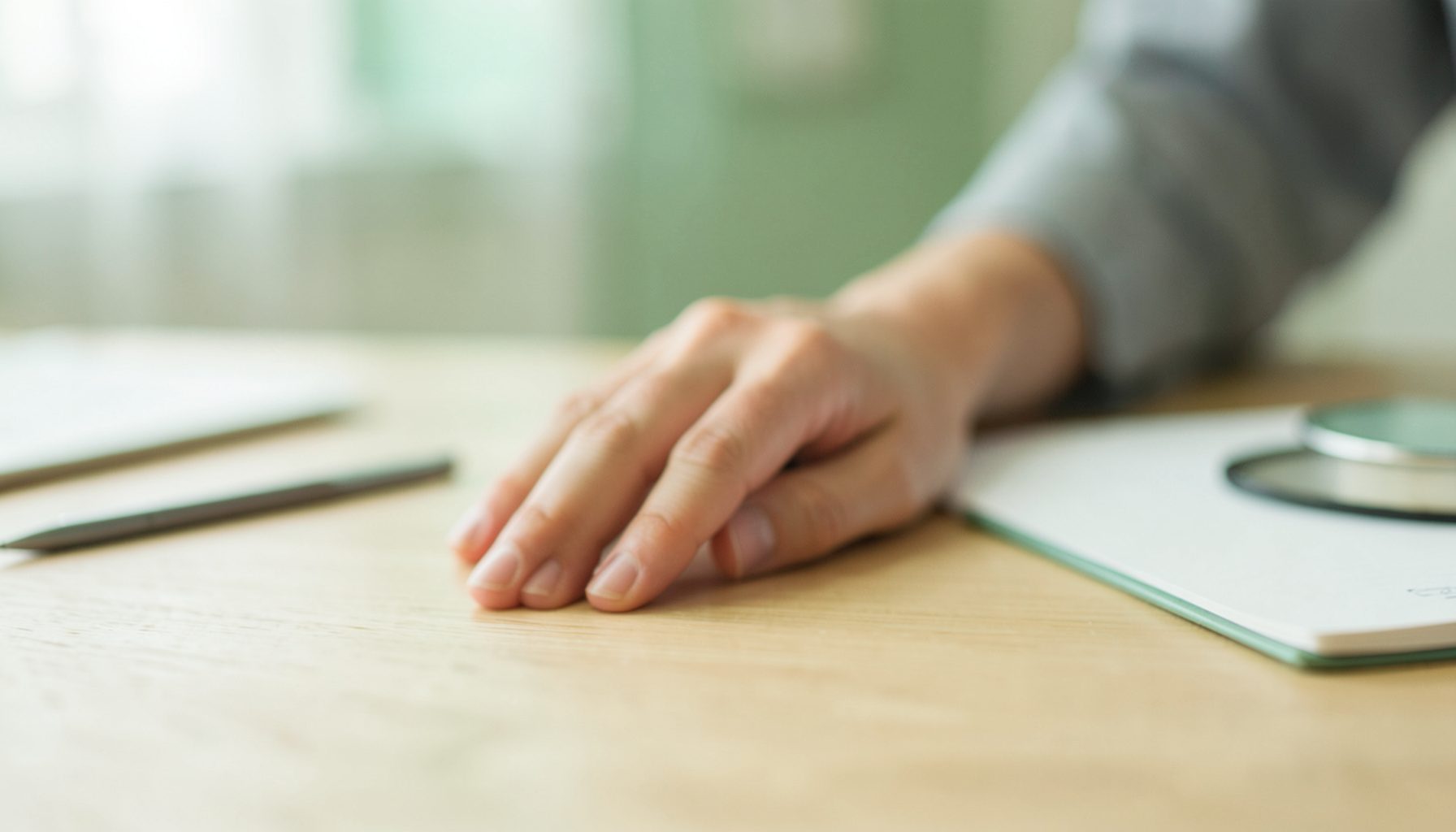 Gros plan flou d'une main posée sur un bureau en bois clair, à côté d'un stylo et d'un carnet, ambiance calme et médicale, tons verts et beiges doux.